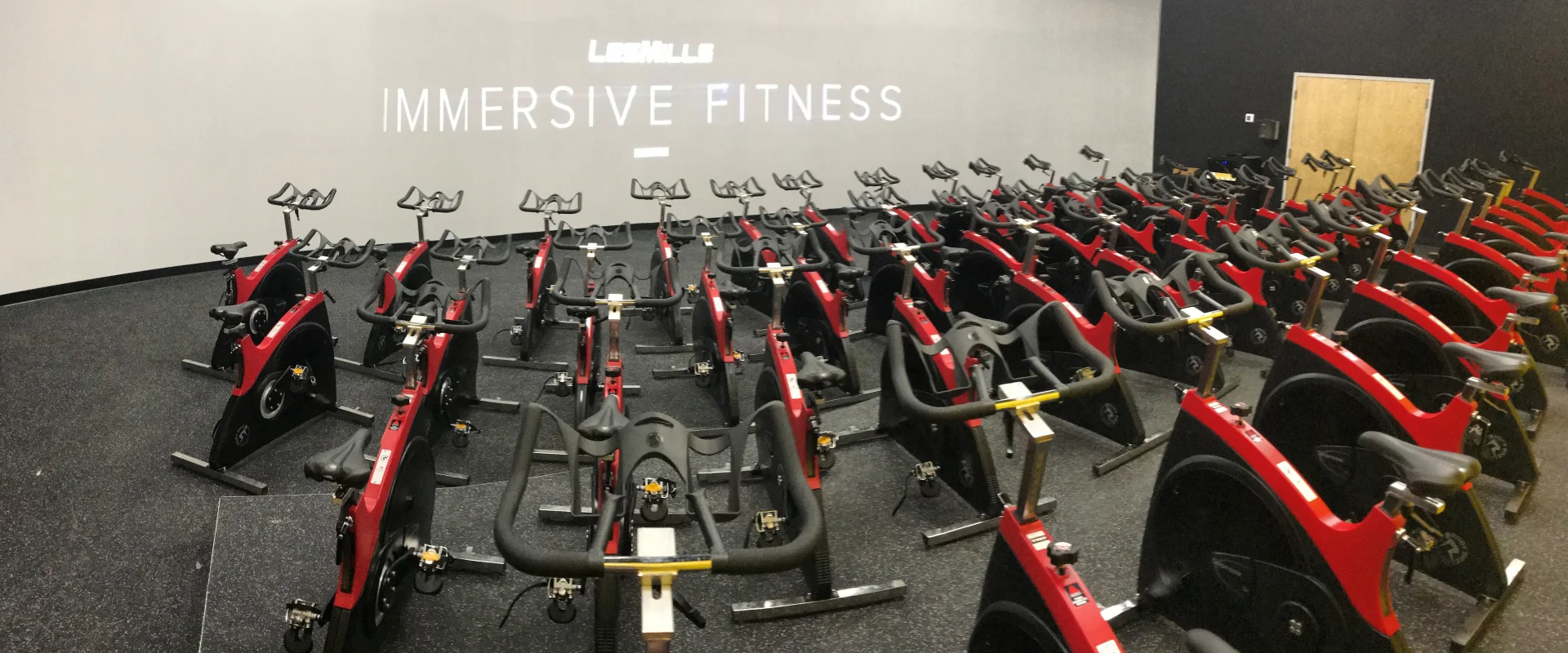 A large room filled with rows of stationary exercise bikes, with the words "Immersive Fitness" displayed prominently on the wall in the background.