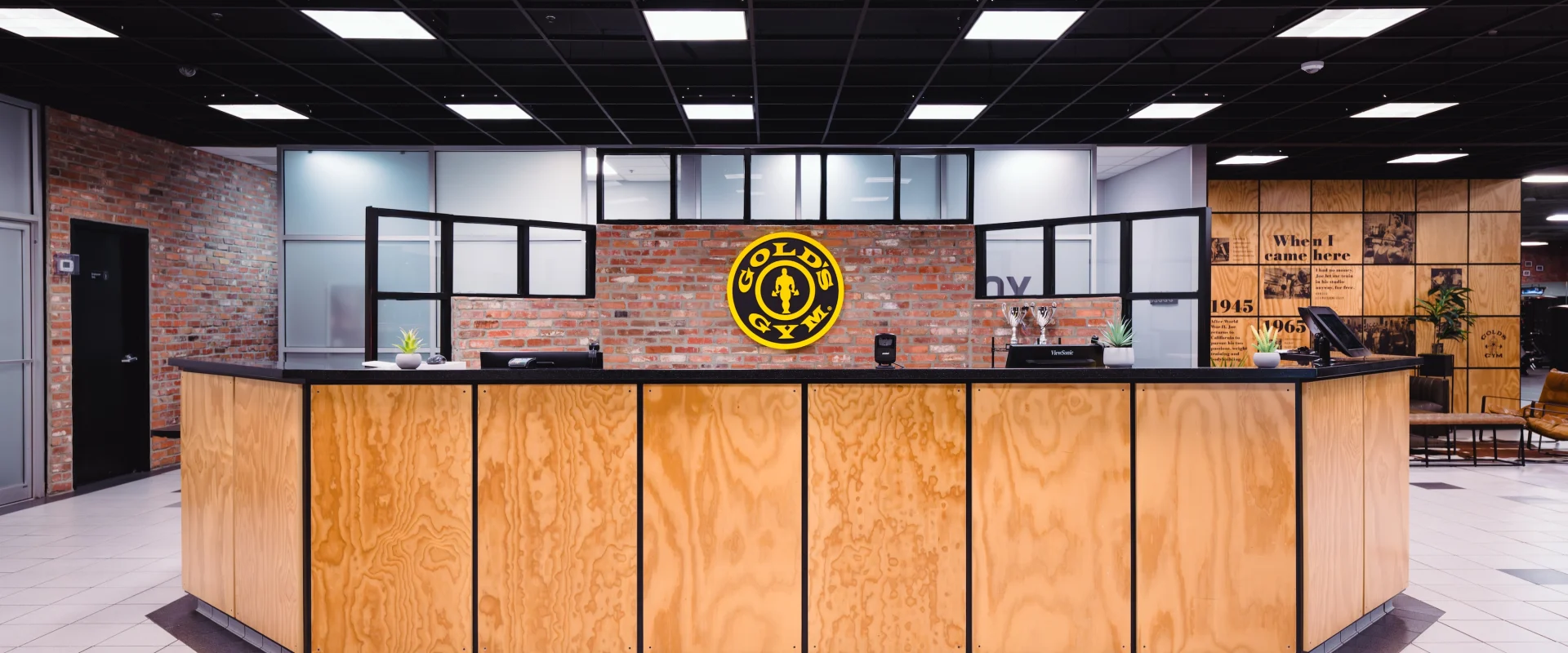 Front desk with a light wood finish, at a gym with a brick wall background and Gold's Gym logo. Modern, welcoming ambiance with potted plants.