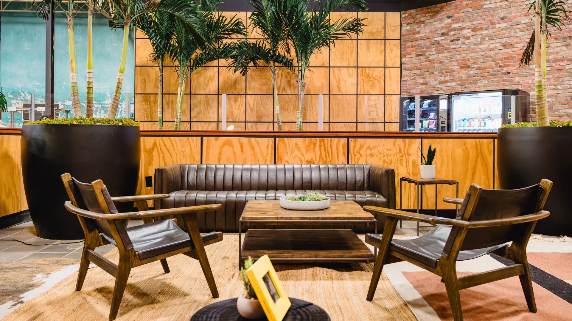 Modern lounge area with a brown leather couch, two wooden chairs, a coffee table, large indoor plants, and a wood-paneled wall with a vending machine in the background.