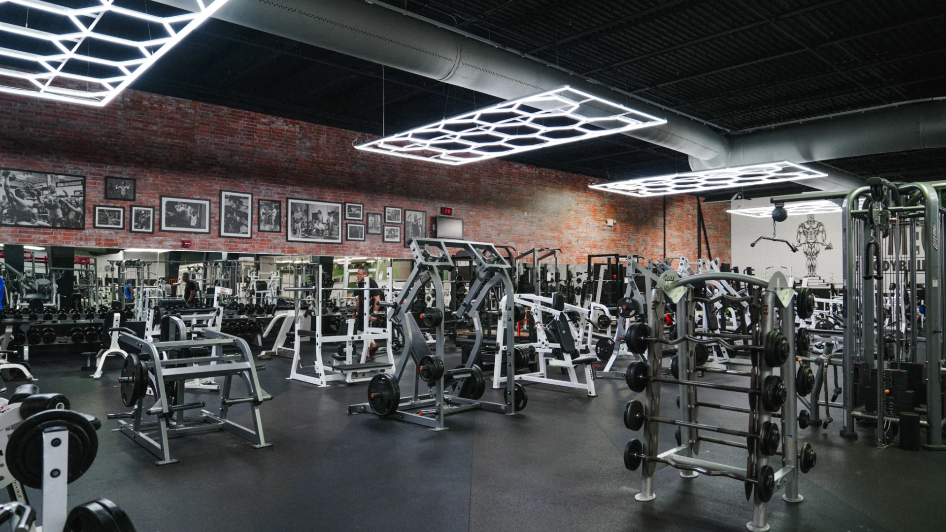 A wide view of Gold's Gym Brassfield's main workout area, filled with plate-loaded and selectorized weight machines. Neon LED lights illuminate the open space, and a red brick accent wall features classic bodybuilding images. The "Mecca of Bodybuilding" mural stands prominently on the far wall.