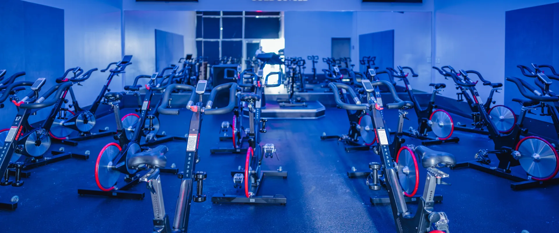 Group Cycle studio at Gold’s Gym in Texas (N. Round Rock), featuring rows of stationary bikes arranged facing an elevated instructor platform. The room is illuminated with blue accent lighting, creating an immersive cycling environment. Wall-mounted TVs are positioned above large mirrors at the front of the room. The Gold’s Cycle logo is displayed on the mirrored wall, and the studio has rubber flooring for durability and support during high-intensity sessions.
