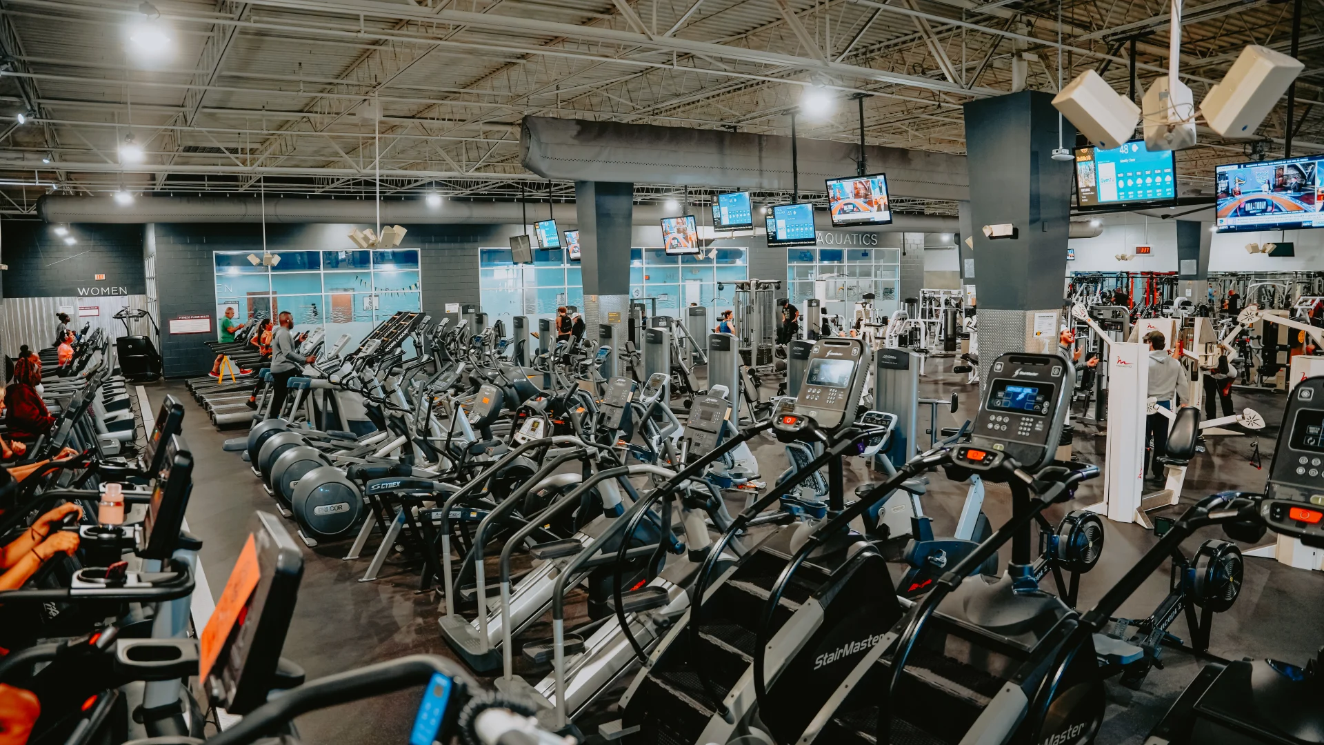 Cardio equipment area at Gold’s Gym Tennessee, Lee Highway, featuring rows of treadmills, elliptical machines, and stair-climbing machines on dark rubber flooring. The space includes a high ceiling with exposed beams, hanging light fixtures, and multiple mounted screens displaying workout stats and entertainment. A view of the aquatics center and machine weight area is visible in the background.