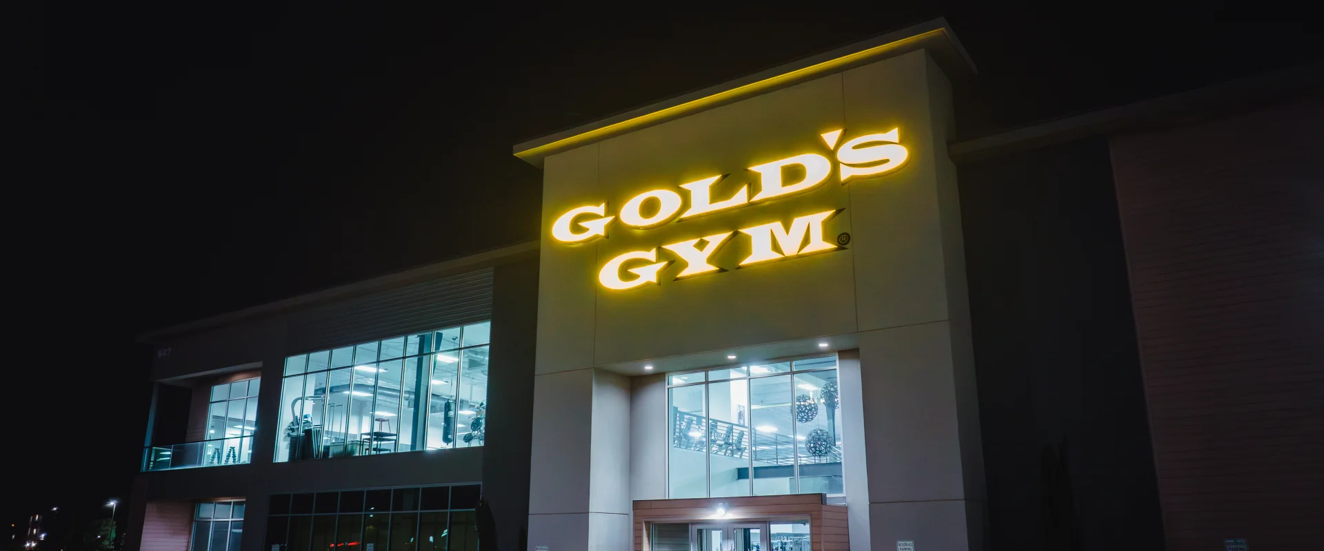 Exterior of Gold's Gym Oklahoma City (Moore) at night, featuring a brightly illuminated Gold's Gym sign above large glass windows showcasing the gym's interior and workout equipment.