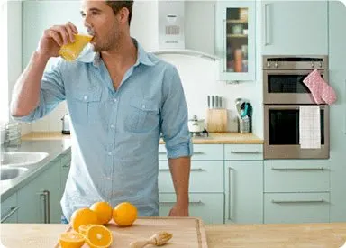 A man in a light blue shirt stands in a kitchen, drinking a glass of orange juice while surrounded by various kitchen appliances and utensils on the countertop.