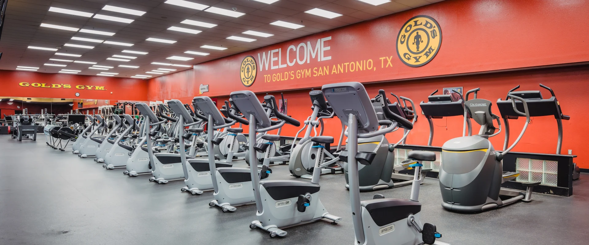 Bright gym interior with rows of modern exercise machines, red walls, and signage reading "Welcome to Gold's Gym San Antonio, TX." Clean and inviting atmosphere.