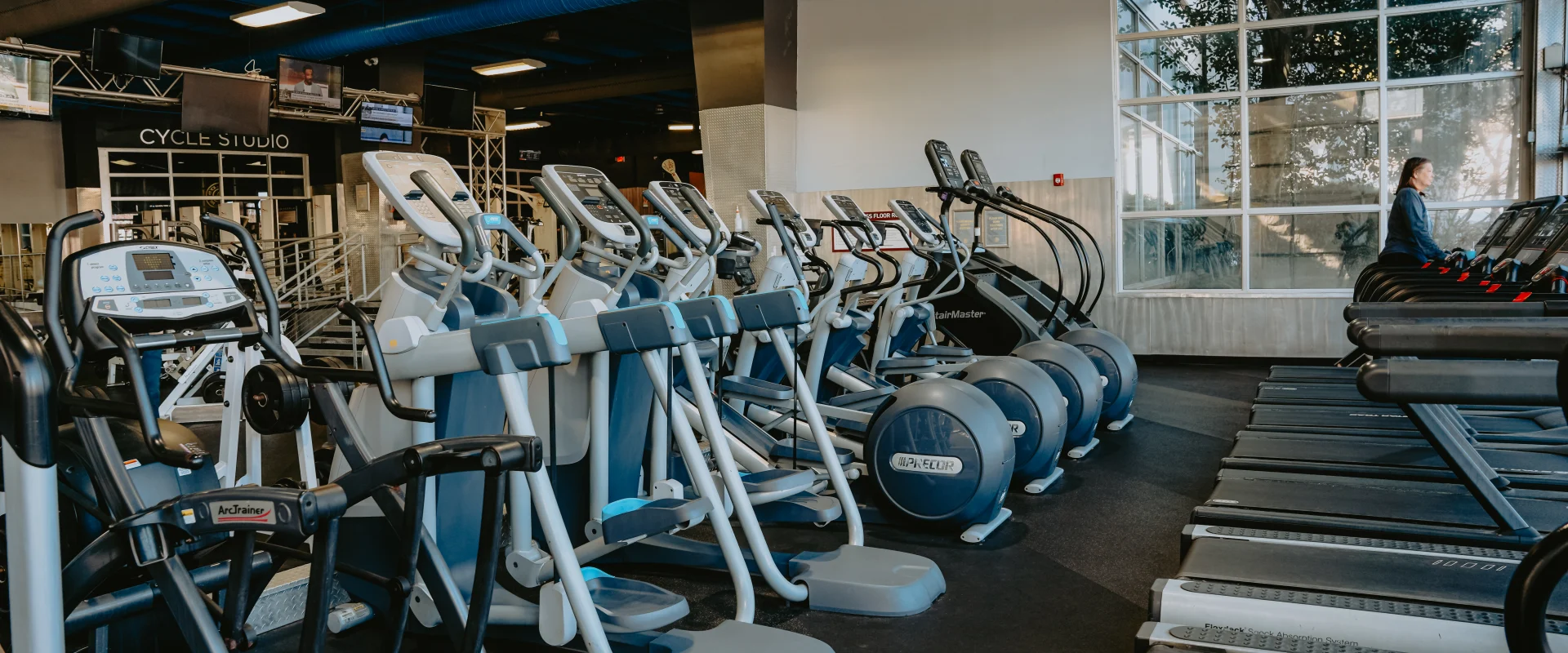 Cardio area at Gold’s Gym Tennessee, Downtown Chattanooga, featuring rows of elliptical machines, treadmills, and an Arc Trainer on a dark rubber floor. The space includes a high ceiling with exposed blue ducts, overhead lighting, and large windows showcasing natural light. A Cycle Studio entrance and mounted televisions are visible in the background.
