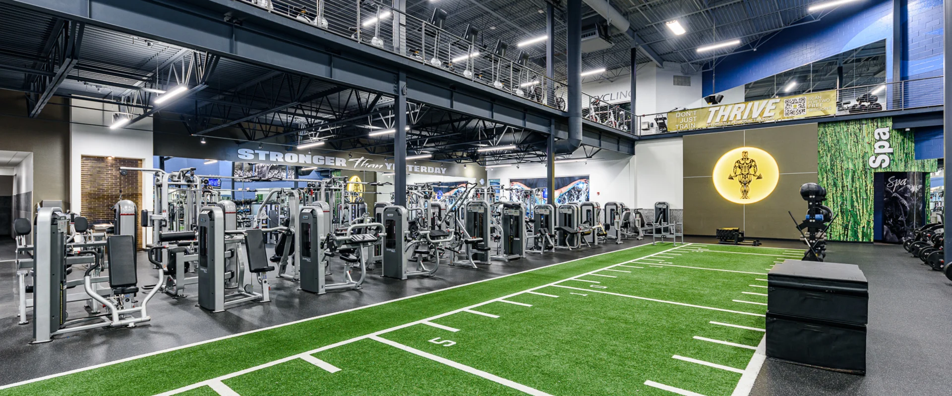 A dynamic functional training area featuring an indoor turf field with white yardage markings. In the background, rows of treadmills and cardio equipment are visible under bright overhead lighting.