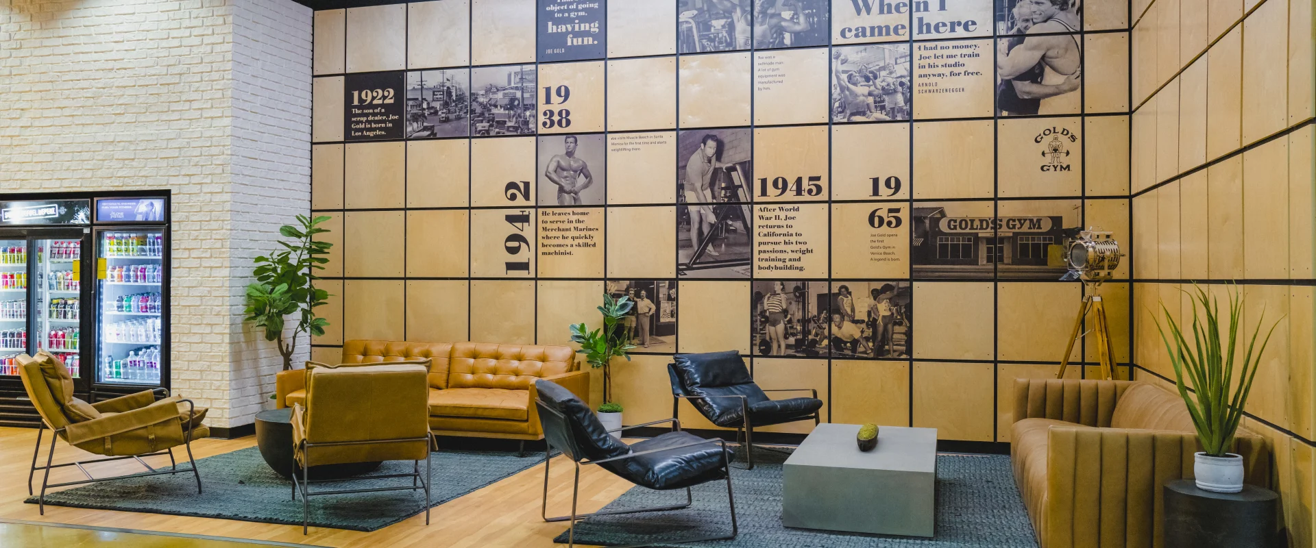 Lounge area at Gold's Gym San Marcos with leather and upholstered seating, a timeline wall showcasing the gym's history, and a nearby refrigerator stocked with beverages.