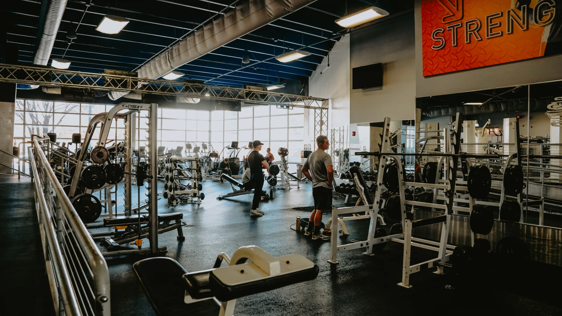 Free weights area at Gold’s Gym Tennessee, Downtown Chattanooga, showcasing weight benches, racks of weight plates, and a Smith machine on a dark rubber floor. The space features large windows allowing natural light, a high ceiling with exposed blue ducts, and overhead lighting. Gym members are visible utilizing the equipment, with additional workout areas and a "Strength" motivational banner in the background.