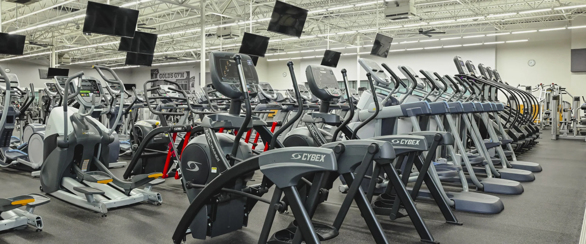 Cardio area at Gold's Gym College Station (Tower Point), featuring rows of treadmills, ellipticals, and stationary bikes, with multiple overhead TVs and bright industrial lighting.