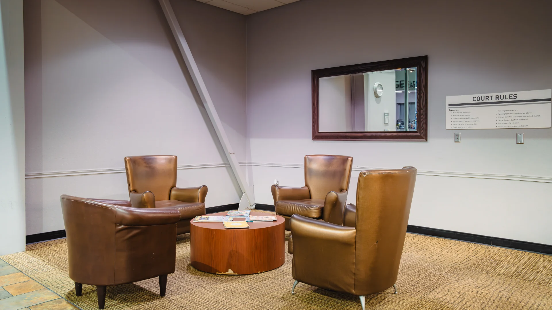 Lounge area in Gold's Gym Waco featuring three brown leather chairs around a round wooden table, set on a tan carpet, with a large wall mirror and court rules signage in the background.