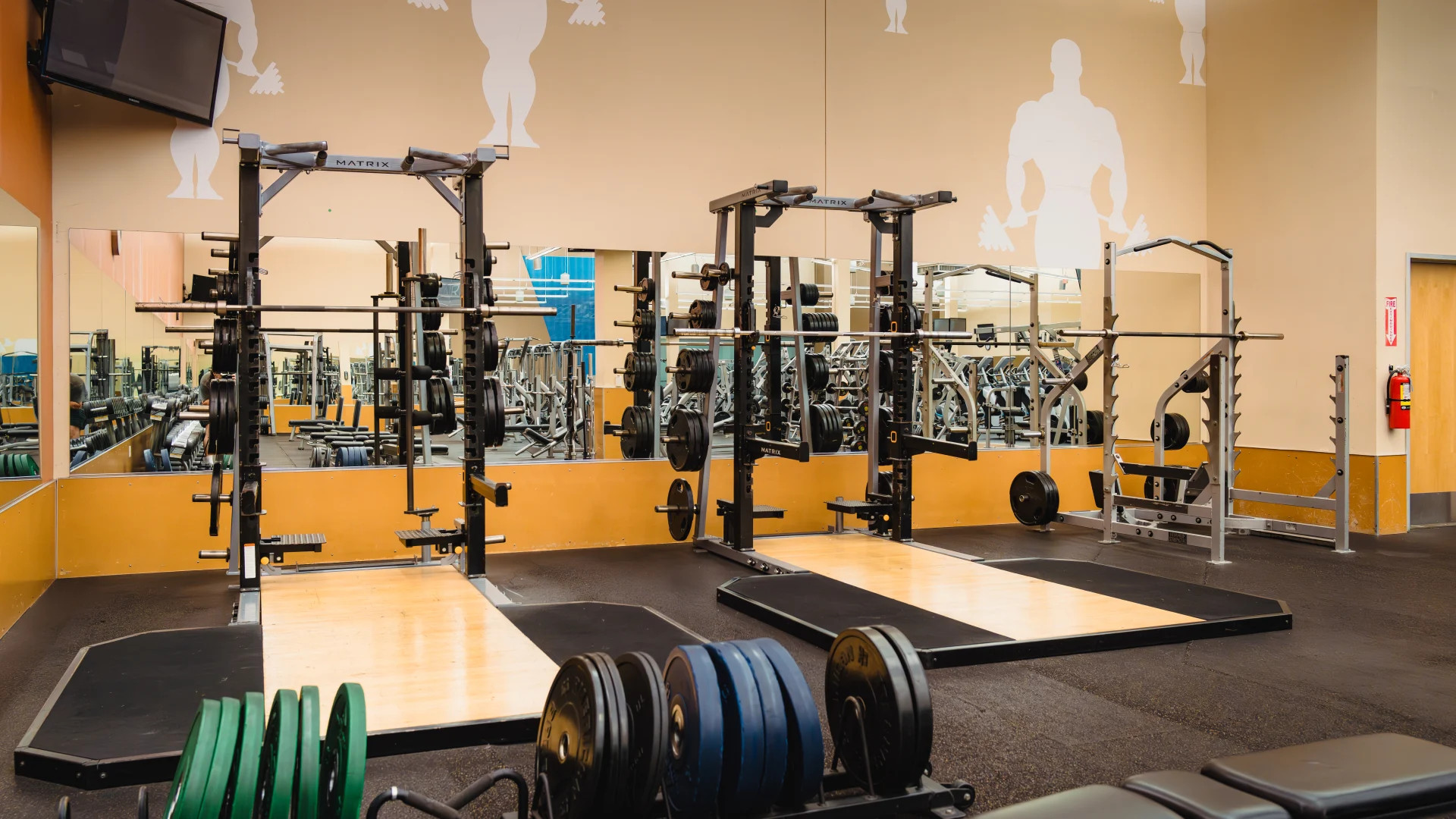 Olympic lifting platforms with two racks holding barbells and weight plates, positioned on wood and rubber surfaces. Green and black weight plates are stacked on a nearby storage rack. A mounted TV and silhouette wall designs of weightlifters are visible above, with large mirrors reflecting gym equipment.