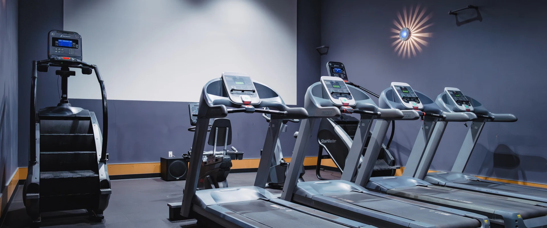 Gym room with a dimly lit ambiance, featuring three treadmills in a row and a stair stepper to the left. A decorative light fixture is on the wall.