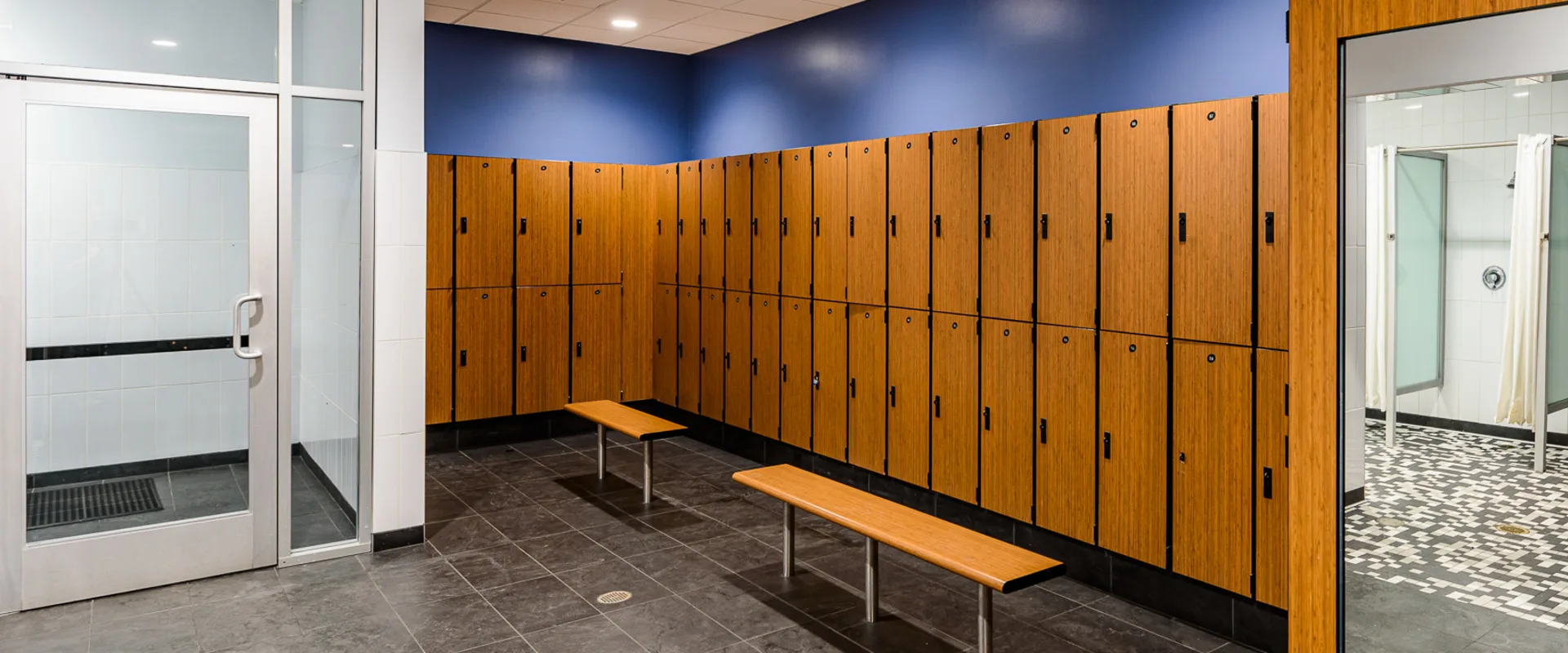 A modern gym locker room with wooden lockers lining the walls, black tiled flooring, and wooden benches for seating. A glass door leads to a steam room, and an open doorway reveals a tiled shower area with frosted glass partitions and privacy curtains.