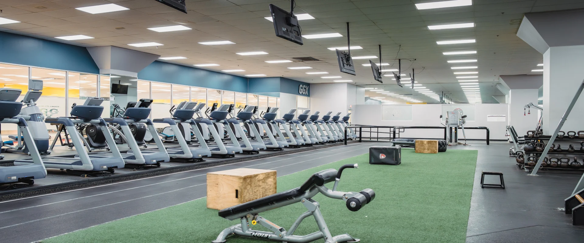Functional training area at Gold’s Gym in Austin (South Central), featuring green turf flooring with equipment such as a decline bench, plyometric boxes, and weighted bags. The space includes kettlebell racks and parallel bars for bodyweight exercises. Adjacent to the turf, rows of treadmills face mounted TVs, with bright overhead lighting creating an open, energetic environment.