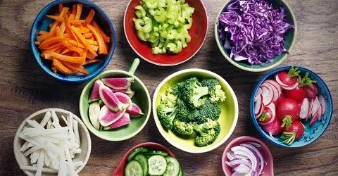 An assortment of colorful, freshly prepared vegetables arranged in various bowls, including carrots, celery, broccoli, radishes, and purple cabbage, set against a dark background.