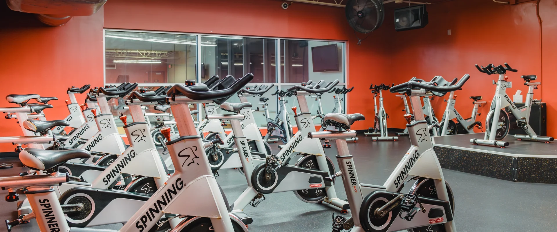 Cycle studio at Gold's Gym Fiesta Trails, featuring rows of stationary bikes, shaded windows, rubber padded floor and bright red accents.