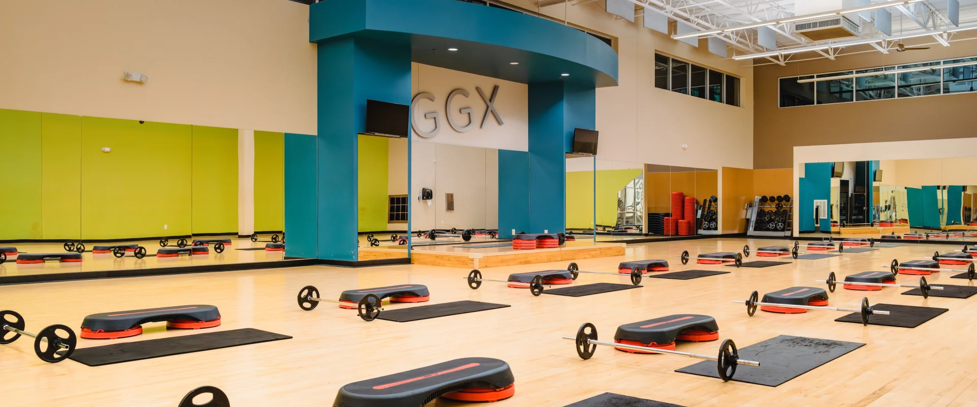 Spacious gym studio with bright wood floors, lined with black mats, red step platforms, and barbells. Large mirrors and vibrant wall colors add energy.