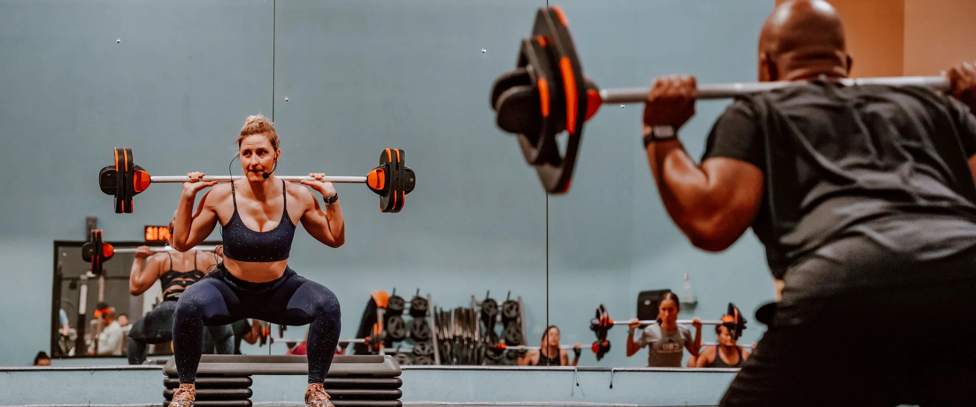 A group fitness class at Gold's Gym Smyrna, Tennessee, featuring a woman leading a barbell squat on an elevated platform, wearing a microphone headset. Other participants are visible in the mirror reflection performing the same exercise with barbells. The room has light blue walls, fitness equipment racks, and a mirrored wall.
