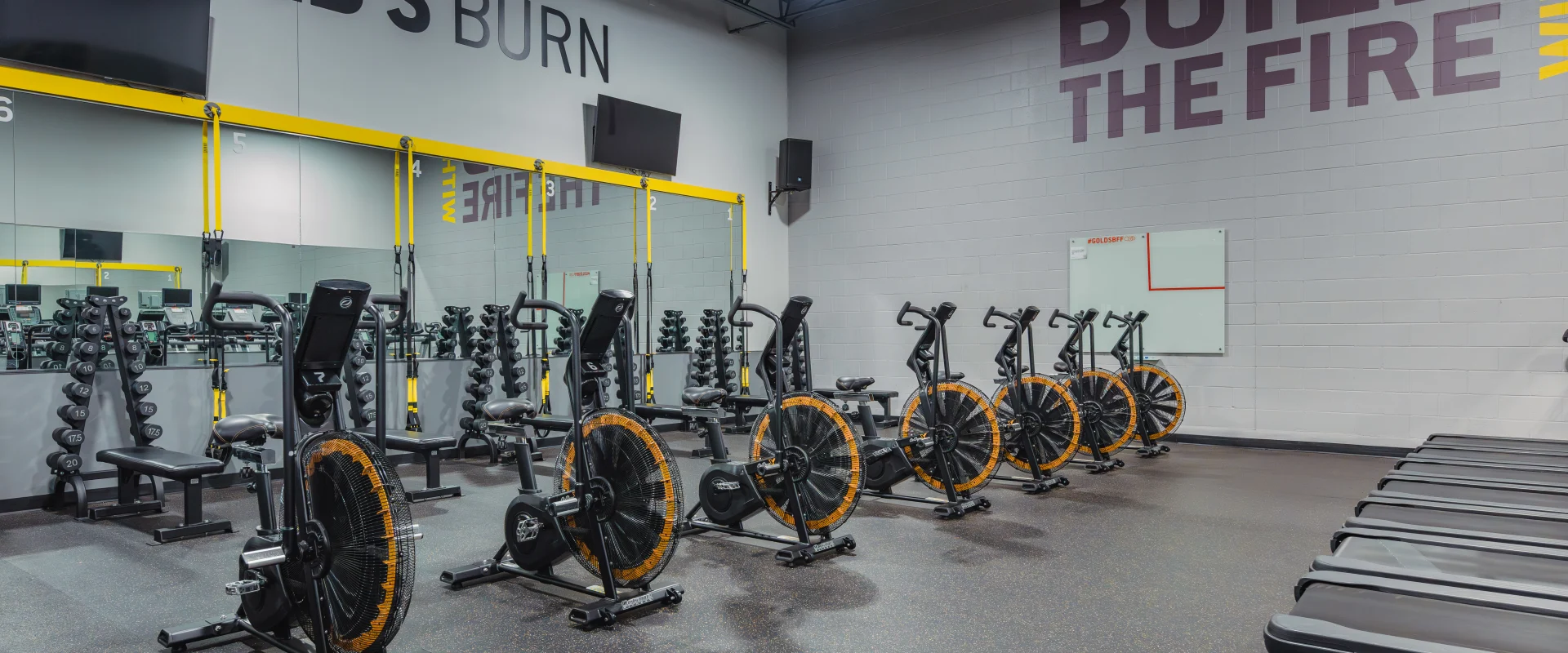 Row of assault bikes with treadmills in front, located in the Gold's Burn section of the gym, surrounded by dumbbell racks, mirrors, and motivational wall text reading "Build the Fire Within.