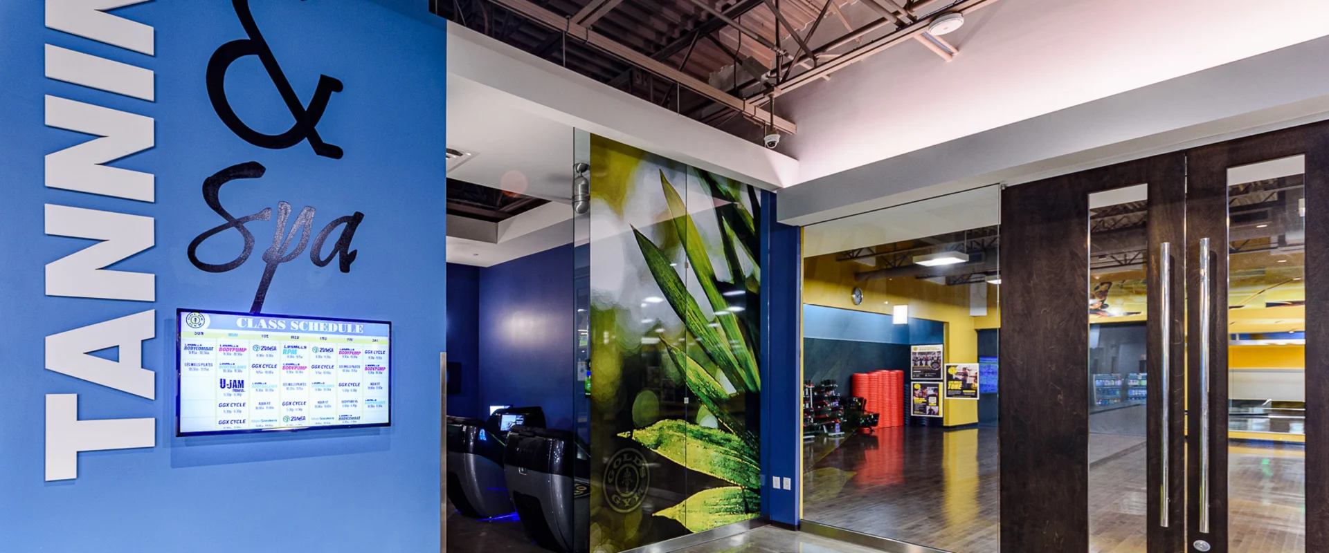 Entrance to the spa and tanning section at Gold’s Gym Garden Grove, featuring a bold blue wall with “Tanning & Spa” in large stylized lettering. A digital class schedule display screen is mounted nearby. Glass doors lead to the group exercise studio, while a modern aloe vera-themed mural adds a wellness-inspired touch.