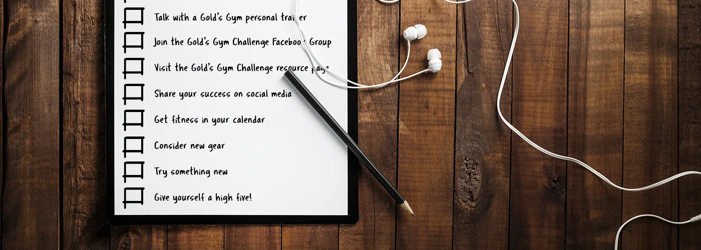 A checklist on a framed whiteboard with various tasks, including "Talk with a Goals Guy personal trainer" and "Join the Goals Guys Challenge Facebook Group", sits on a wooden surface alongside a pair of earphones.