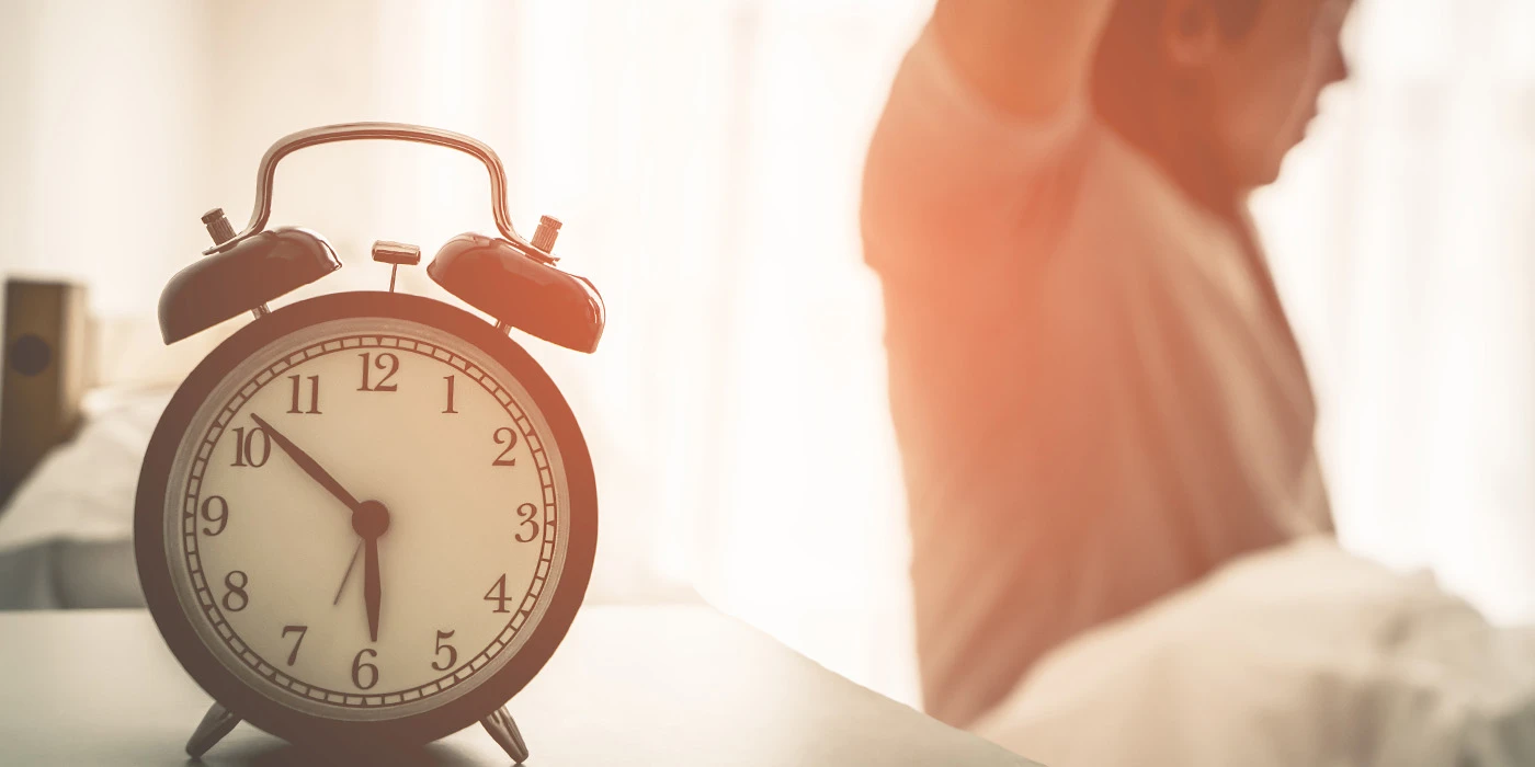 A vintage-style alarm clock sits on a surface, with a blurred curtain or fabric in the background.