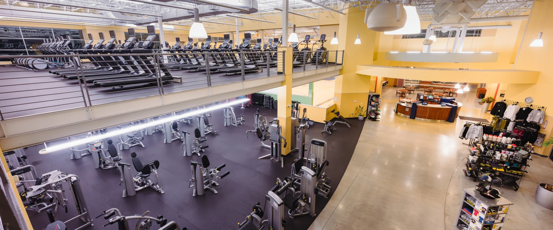 Spacious gym interior with cardio machines on upper level, strength equipment below. Bright lighting and yellow walls create an energetic atmosphere.