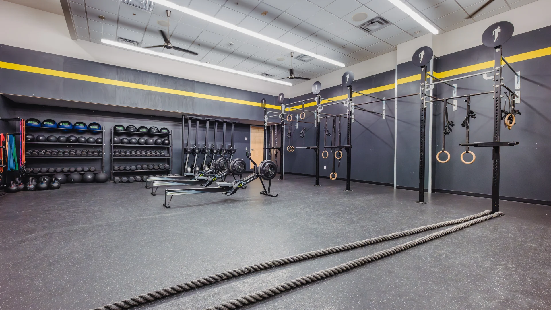 Gold's Fit® room at Gold's Gym Austin (Downtown) featuring a functional training space with dark walls, yellow accents, and ceiling fans. The room includes pull-up rigs, gymnastic rings, battle ropes, rowing machines, and shelves stocked with kettlebells and medicine balls, creating a focused workout environment.