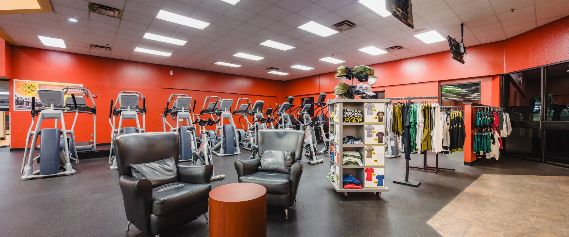 A gym interior featuring rows of elliptical machines against a red wall. In the foreground, two black chairs and a table are near a display of gym apparel.