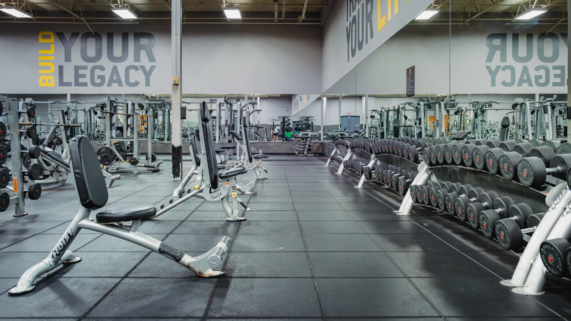 Weightlifting area at Gold's Gym Waco featuring rows of dumbbells on sleek racks, adjustable benches on black rubber flooring, mirrored walls reflecting the equipment, and motivational wall text reading "Build Your Legacy" and "Transform Your Life."