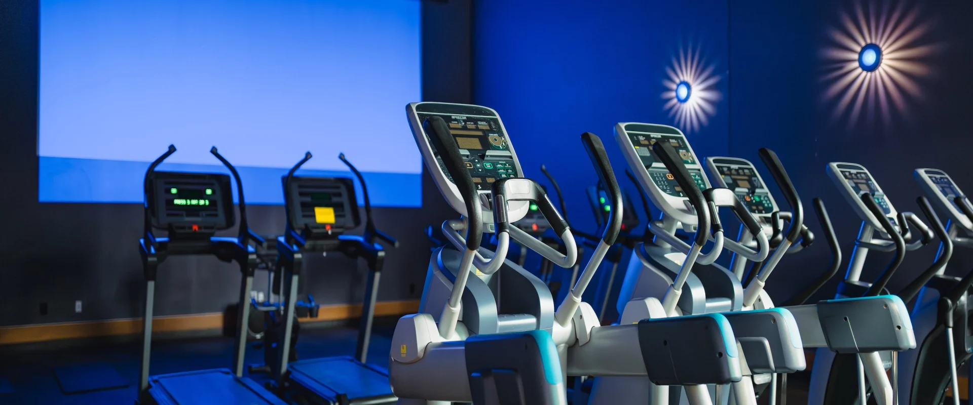 Cardio Cinema at Gold’s Gym in Austin (Westlake), showcasing rows of treadmills and ellipticals facing a large projection screen. The dim, blue-tinted lighting and wall sconces create a theater-like atmosphere, enhancing the workout experience with immersive visuals. The equipment features digital consoles for personalized cardio tracking.