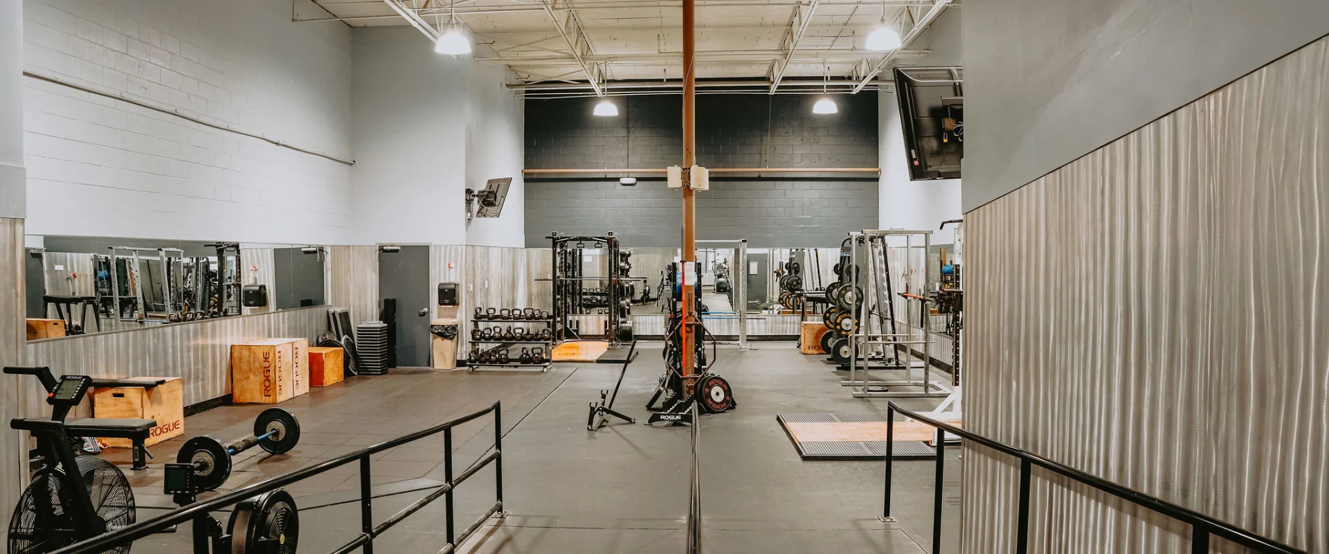 Circuit training area at Gold’s Gym Tennessee, Hixson, featuring a variety of equipment, including a rowing machine, stationary bike, free weights, and plyometric boxes, all set on a rubberized floor. The room has corrugated metal accent walls and mirrored panels reflecting the equipment, adding depth to the space. High ceilings with exposed white ducts and industrial lighting create a bright, open atmosphere. A wall-mounted TV, training racks, and barbells are visible in the background, providing a functional and versatile workout environment.