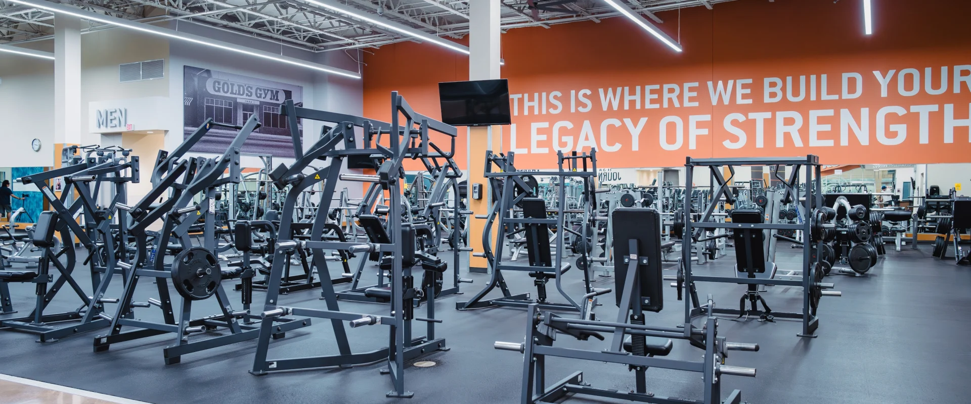 A spacious gym interior with various weight machines on a clean, dark floor. An orange wall reads, “This is where we build your legacy of strength.”