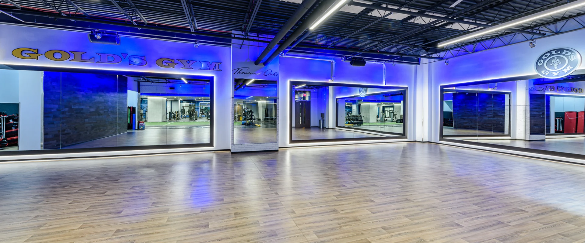 Group exercise studio at Gold’s Gym Thousand Oaks, featuring a spacious wood-floored area with full-length mirrors along the walls. Soft blue LED lighting creates a modern atmosphere. The open layout accommodates a variety of fitness classes, with ceiling fans and an advanced sound system enhancing the workout experience.