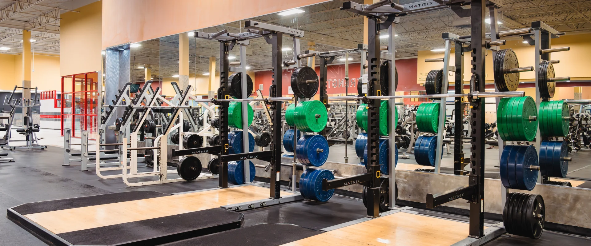 Racks of colorful weight plates and barbells on lifting platforms. Brightly lit, with mirrors reflecting exercise equipment.
