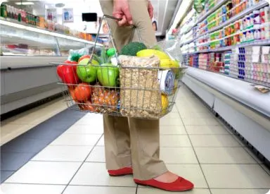 A person's legs and feet are visible in the foreground, wearing beige pants and red shoes, while a shopping basket filled with various fruits and vegetables is held in front of them in a grocery store aisle.