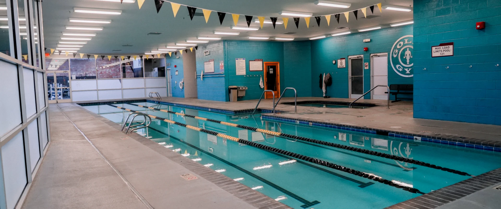 An indoor lap pool at Gold's Gym Brassfield, surrounded by bright turquoise walls and a clean tile deck. Black and gold pennant flags hang from the ceiling, while large windows let in natural light. The pool is equipped with handrails and floating lane dividers for structured swim training.