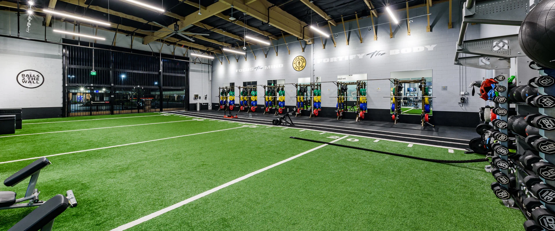 The turf area at Gold’s Gym Cerritos features a large stretch of artificial turf marked with white lines for agility and functional training. A row of squat racks with bumper plates in red, blue, green, and yellow lines the back wall, with mirrors reflecting the space. A ‘Balls to the Wall’ target is painted on one side, while motivational phrases like ‘Focus the Mind, Fortify the Body’ are displayed on the walls. Overhead lighting, exposed beams, and a high industrial ceiling complete the open training environment.