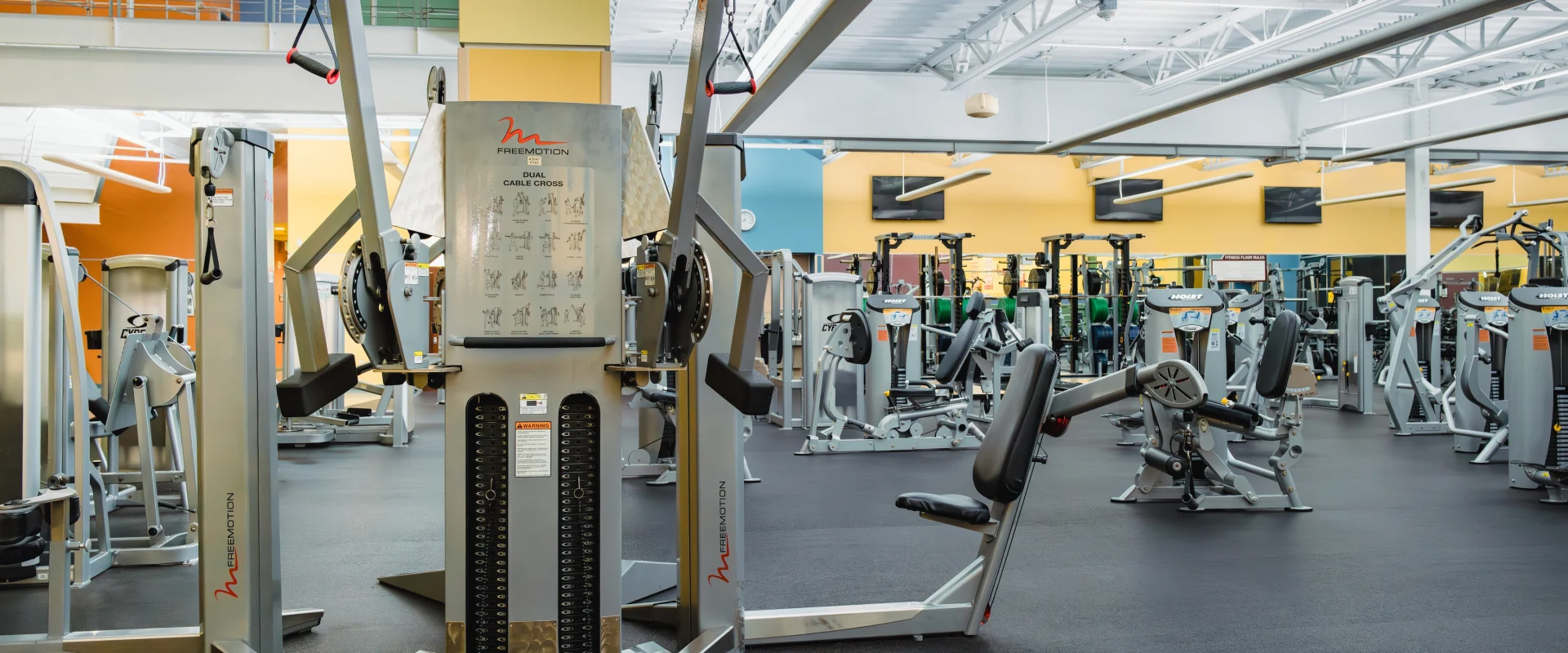 Spacious gym interior with various exercise machines, including a central cable machine. Bright lighting and neutral colors create an energetic atmosphere.