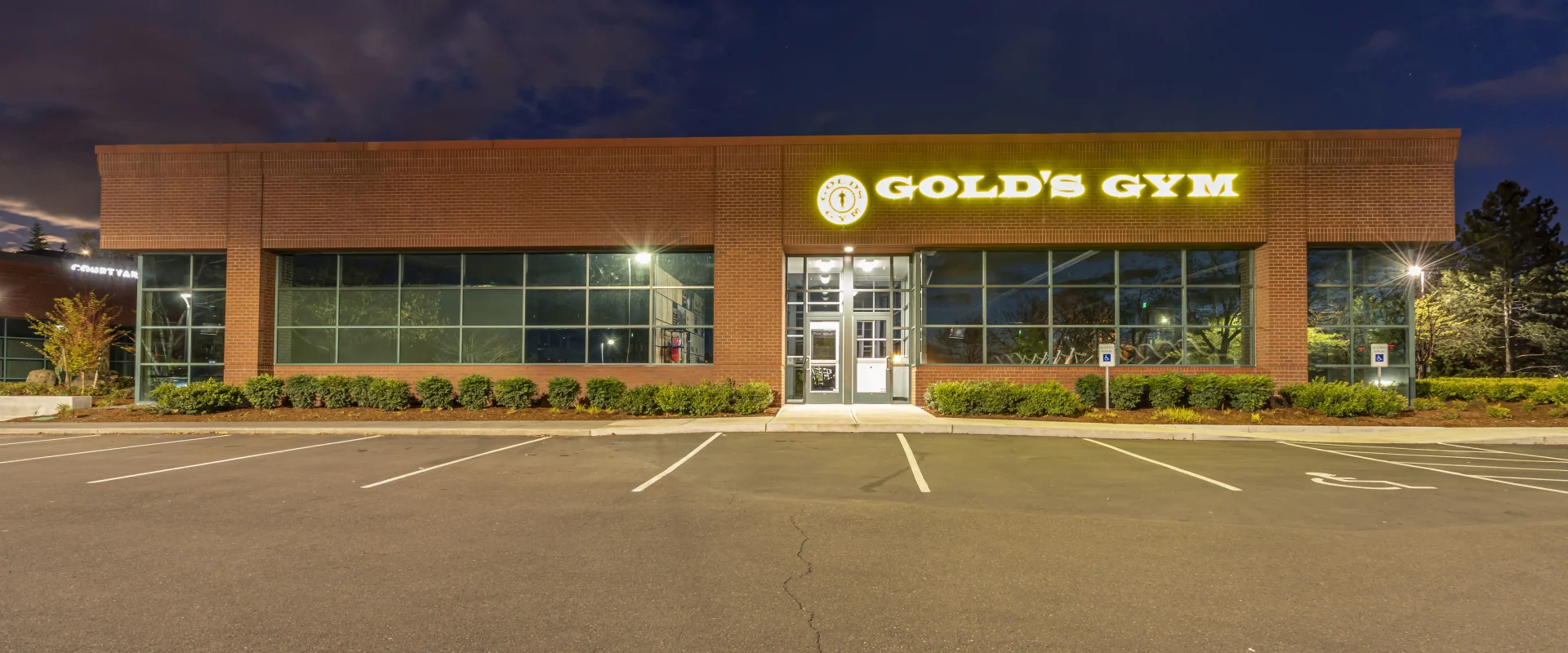A large, well-lit gym building with the name "Gold's Gym" prominently displayed on the facade, surrounded by a paved parking lot in the foreground and a dark, cloudy sky in the background.