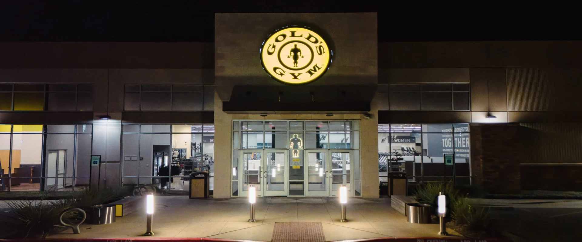Exterior view of Gold's Gym in Texas (Pflugerville), featuring the illuminated Gold's Gym logo above the entrance, large glass doors, and well-lit surroundings with modern light posts and landscaping. The interior gym equipment is visible through the windows, showcasing a welcoming and vibrant facility.