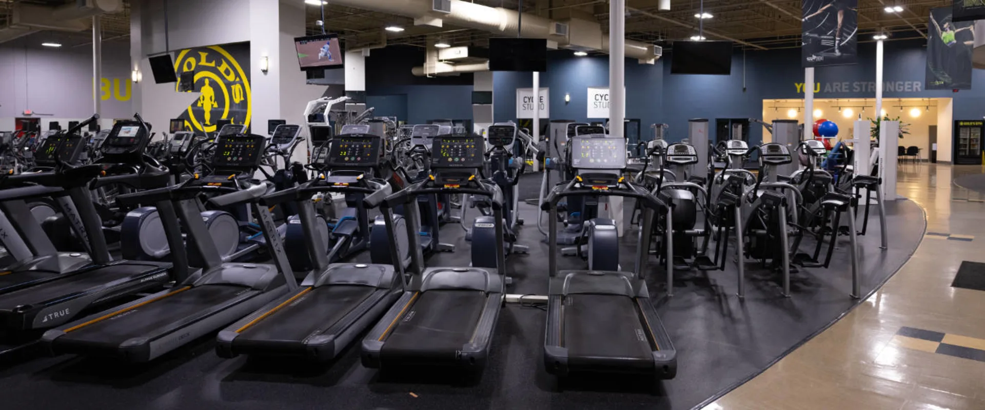 Rows of treadmills, ellipticals, and stationary bikes fill the cardio area at Gold’s Gym in Woodbridge (Lake Ridge). Each machine is equipped with a digital display for tracking workouts. Overhead, several mounted televisions provide entertainment. The Gold’s Gym logo is prominently displayed on a black accent wall in the background. To the right, a section of the gym’s open floor plan reveals additional training areas, including a cycle studio and a lounge space. The flooring transitions from rubberized black to smooth tile near the entrance.