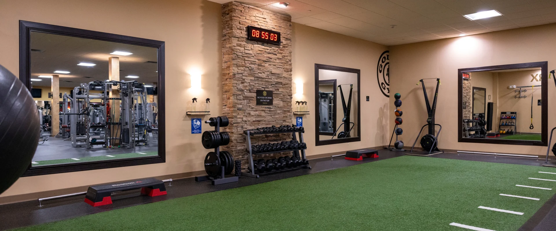 A functional training area in a gym with green turf flooring, white line markings, a stone accent wall with a digital clock, mirrors, dumbbell racks, and neatly arranged gym equipment like medicine balls, kettlebells, and resistance bands.