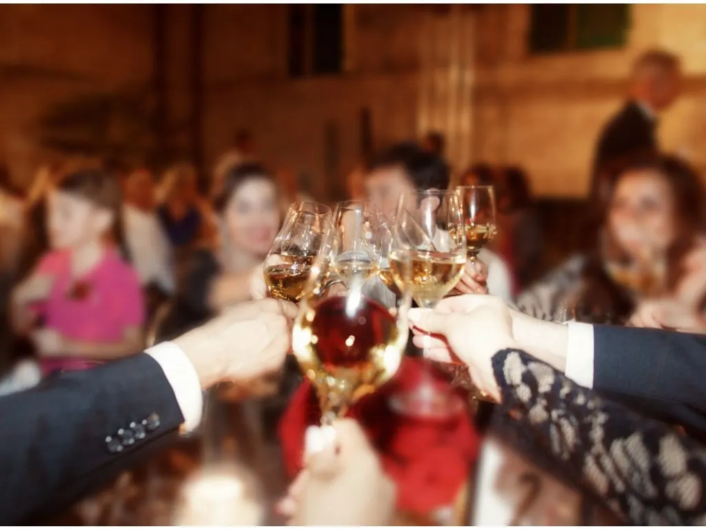 Hands holding up glasses of wine in a crowded, dimly lit room, with people in the background.