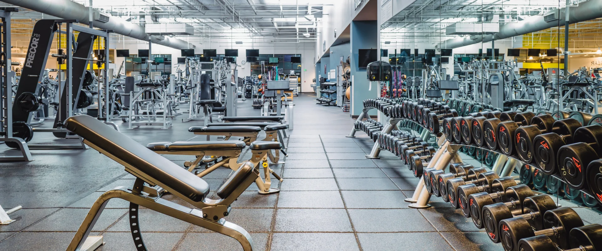A spacious gym with various exercise equipment including benches, dumbbells, and machines. The atmosphere is clean and organized with bright lighting.