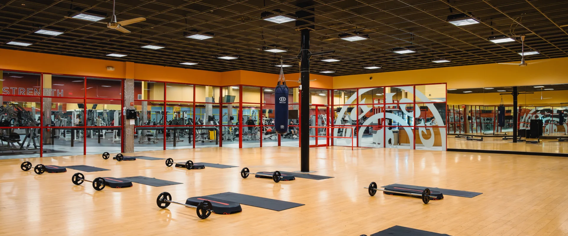 Spacious gym studio with wooden floors, neatly arranged step boards, and weights. Bright lighting, mirrored walls, and a red-accented backdrop create an energetic vibe.