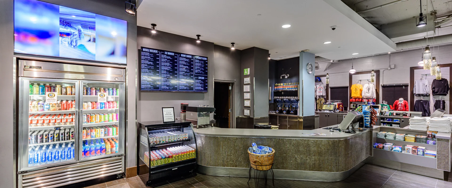 Smoothie bar at Gold’s Gym Hollywood, featuring a curved counter with a tiled front and a cash register. A refrigerated display stocked with bottled beverages stands to the left, with a digital menu mounted above listing smoothie options. A smaller refrigerated case displays protein snacks. Behind the counter, multiple blenders and dispensers are arranged for drink preparation. To the right, gym merchandise, including apparel and towels, is neatly organized on shelves. Pendant lights hang from the exposed ceiling, illuminating the space.