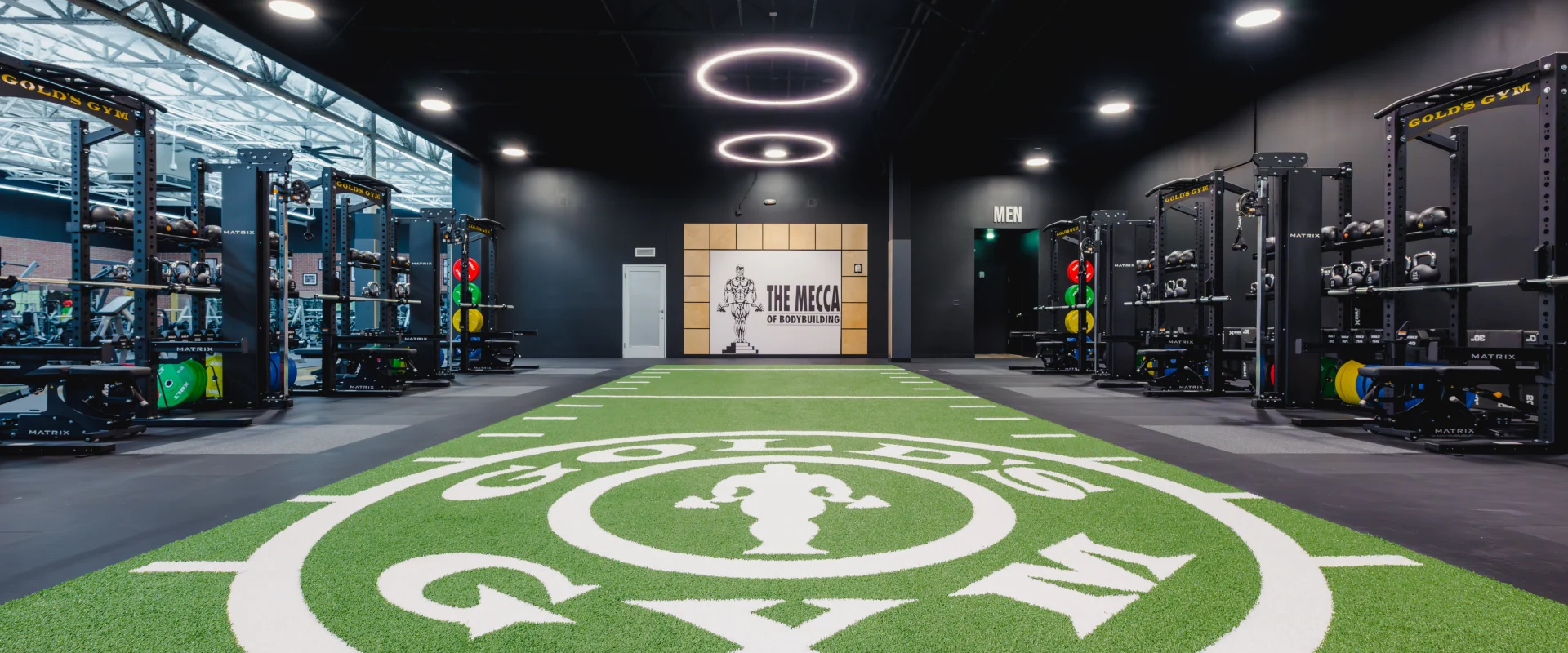Functional training area at Gold's Gym featuring turf with the gym's logo, lined for athletic drills, surrounded by weight racks and illuminated by circular ceiling lights.