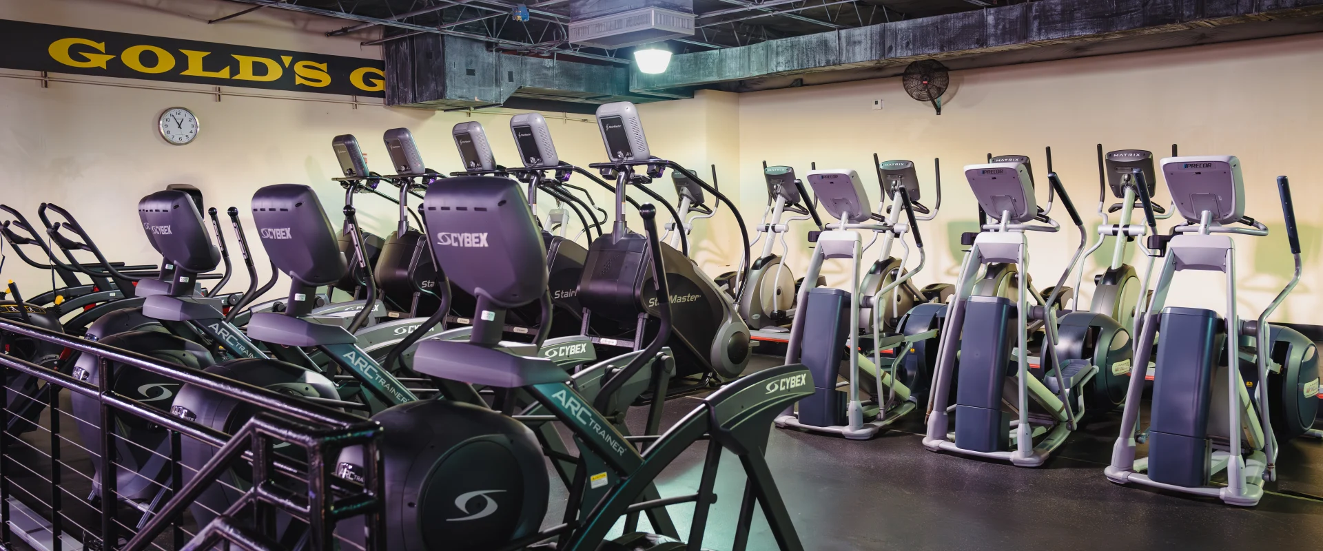 Rows of cardio machines, including elliptical trainers and stair climbers, arranged in a spacious gym area with a light yellow wall and a "Gold's Gym" sign in the background.